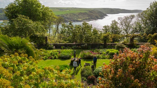 Visitors and a gardener in the terraced gardens with the sea and coastline behind, at Overbeck's, Devon